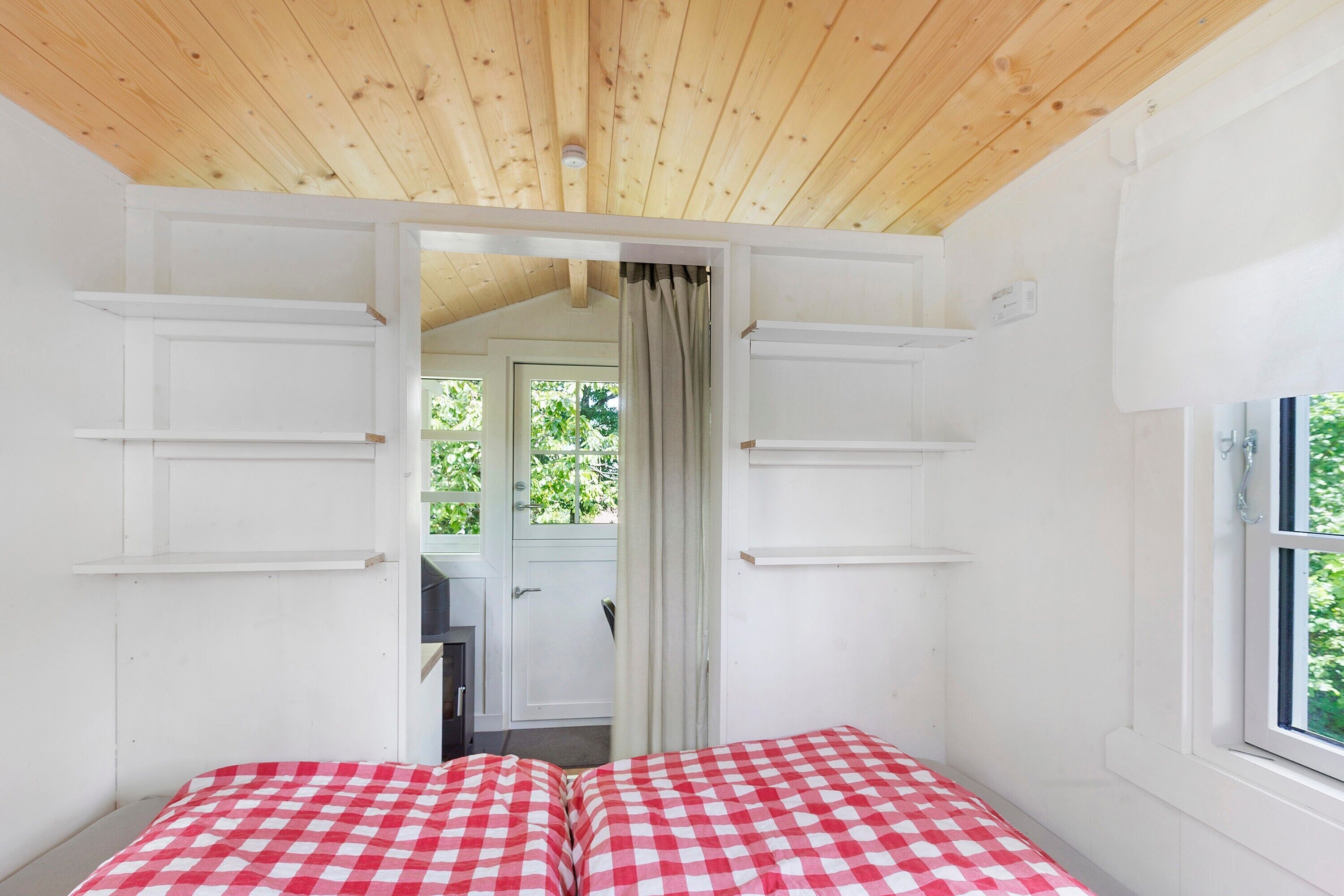 A room with a wooden ceiling, a window with a view of greenery, and a bed with a checkered bedspread.