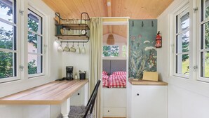 Une pièce de taille modeste, comprenant un bureau en bois, une chaise, un lit avec une literie à carreaux, une étagère fixée au mur avec des ustensiles de cuisine et un extincteur.