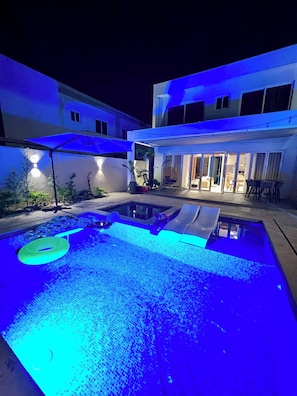 A swimming pool with blue lighting, a covered seating area, and a building in the background. - Luxury Private Villa Pool & Jacuzzi Juan Dolio (Guayacanes)