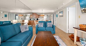 Ein modernes Wohnzimmer mit blauen Sofas, einem Holztisch und einer Küche im Hintergrund.