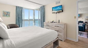 Ein Schlafzimmer mit einem großen Bett, einem Schminktisch, einem Fernseher an der Wand und Blick aufs Meer.