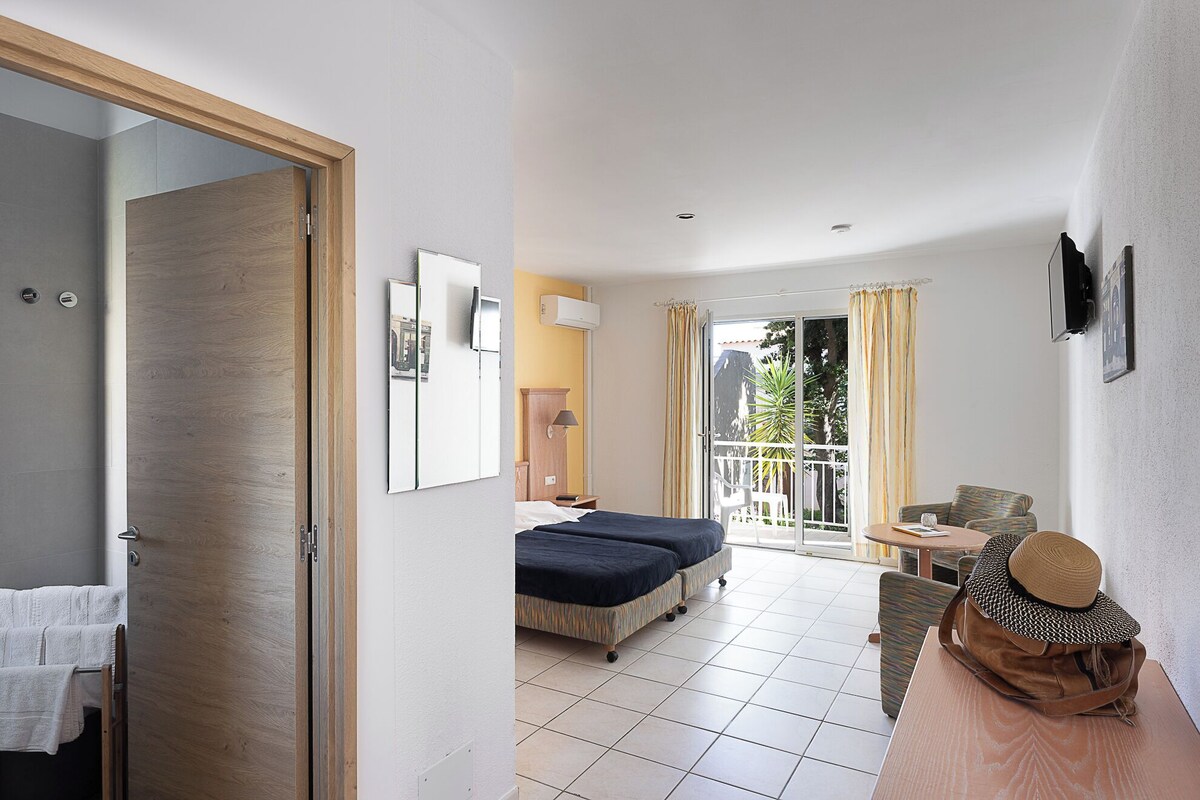 Habitación de hotel con cama, escritorio, una silla, balcón con cortinas y vista al exterior.