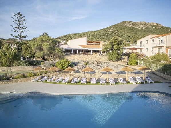 Una piscina con sillones de playa y sombrillas, rodeada de edificios y un paisaje montañoso.