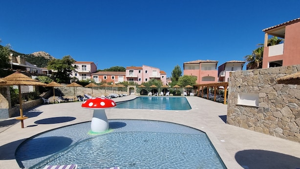 A pool area with lounge chairs, a mushroom-shaped water feature, and buildings in the background. - La Residence Maristella (Aregno)