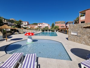 Un área de piscina con sillones reclinables, una fuente con forma de hongo y edificios al fondo.