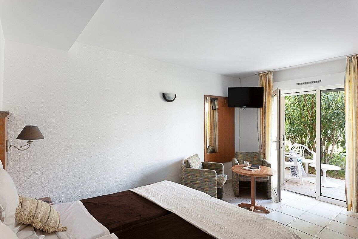 Habitación de hotel con cama, una silla, una mesita, televisor y puerta corrediza de vidrio que da a un área exterior con sillas.