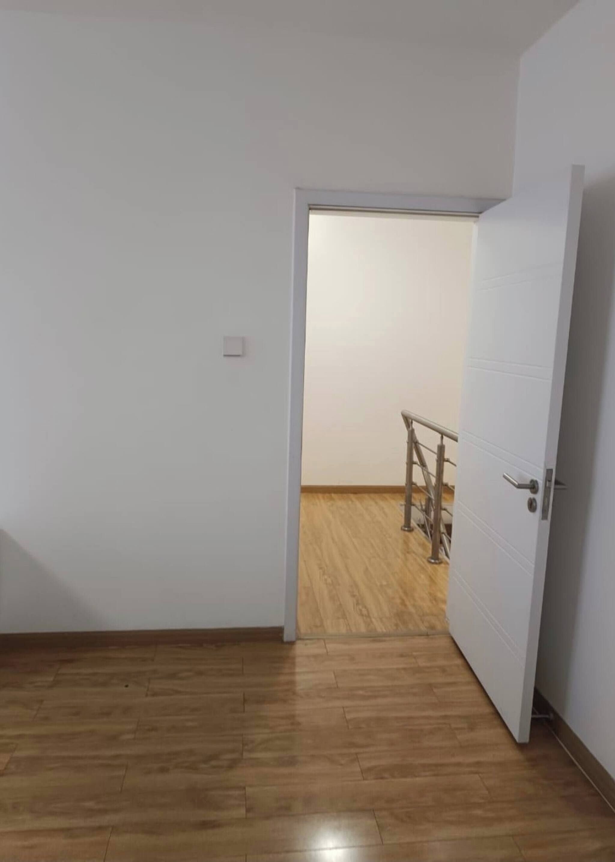 Una habitación con piso de madera, paredes blancas, una puerta y una barandilla metálica al fondo.