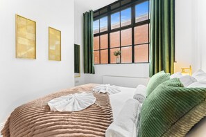 Ein ordentlich eingerichtetes Schlafzimmer mit einem großen Fenster, einem Heizkörper und zwei gerahmten Kunstwerken an der Wand.