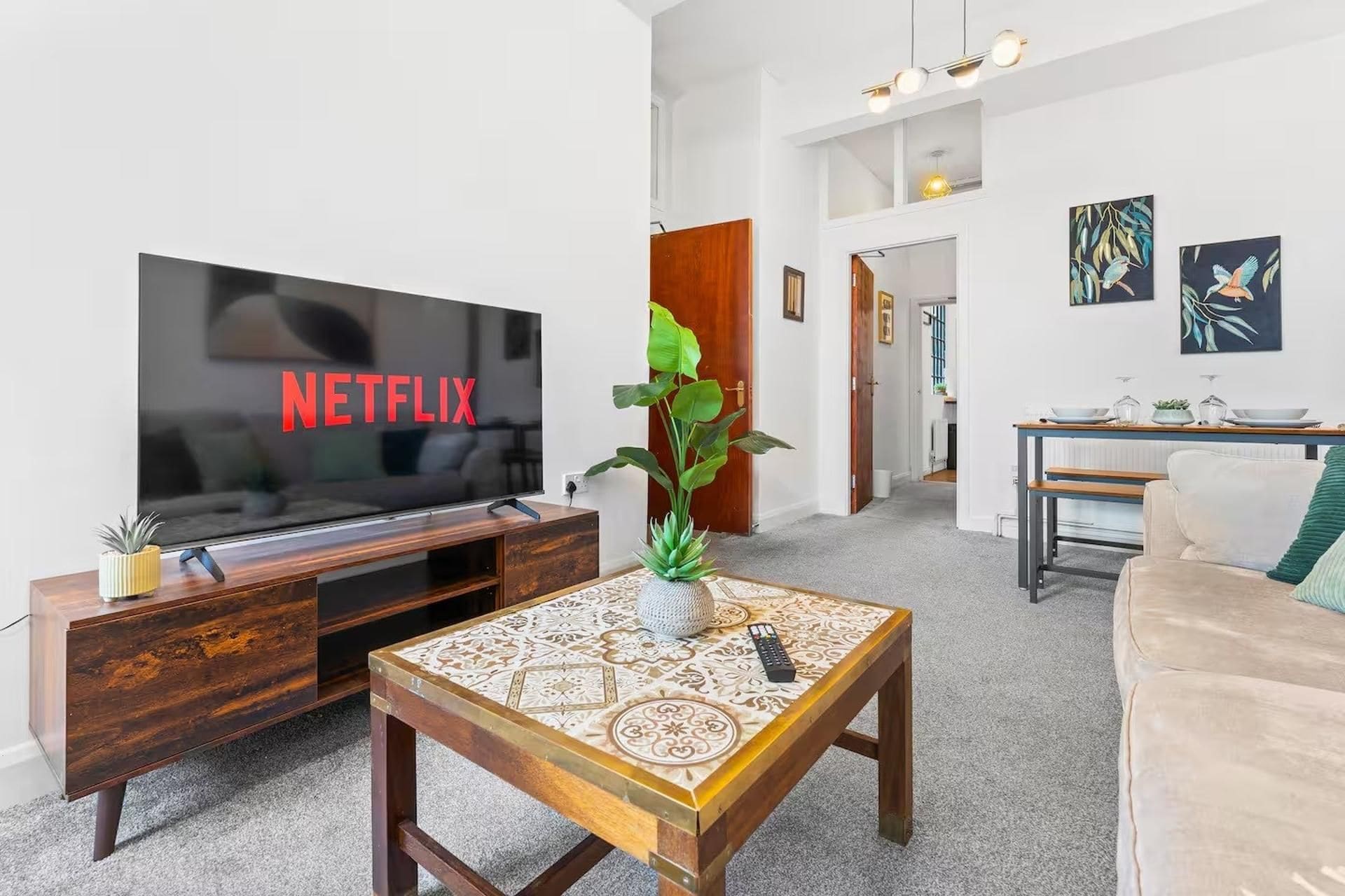 Una sala de estar con una televisión mostrando 'NETFLIX', una mesa de centro con superficie decorativa, un sofá y un comedor con mesa y sillas.