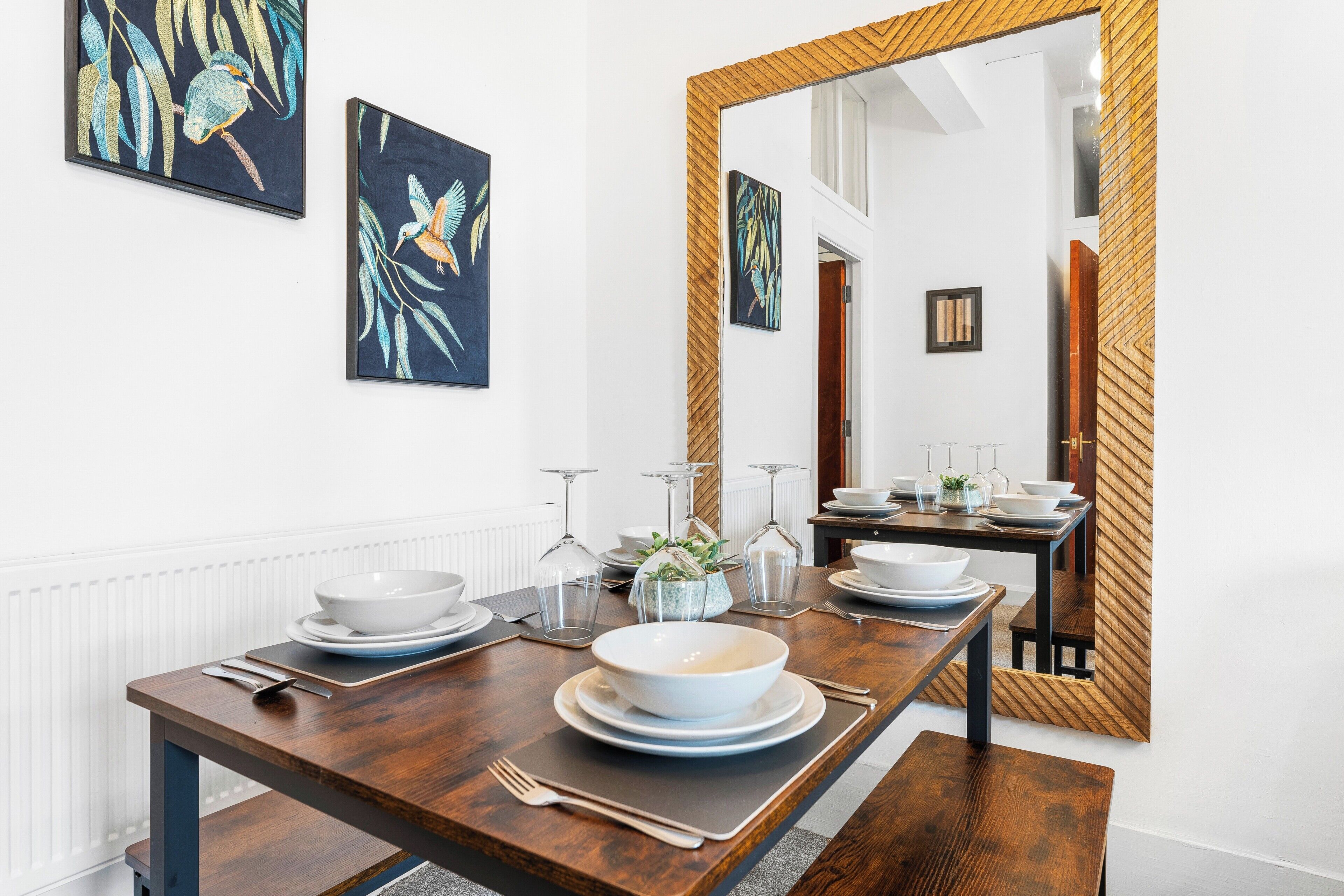 A dining area with a wooden table set for four, a large mirror, and two wall-mounted paintings.
