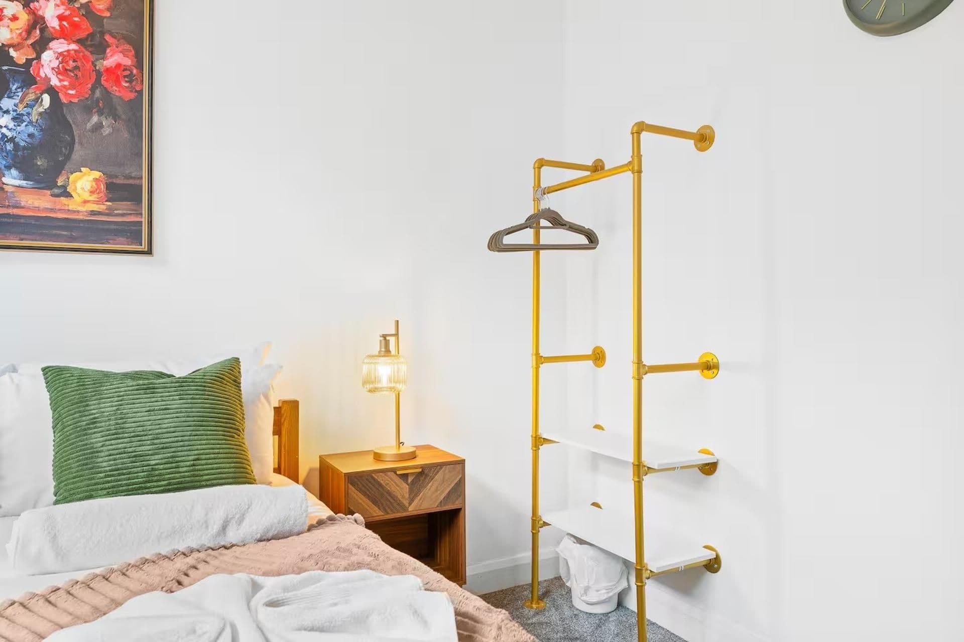 A bedroom with a bed, a bedside table with a lamp, a hanging clothes rack, and a painting on the wall.