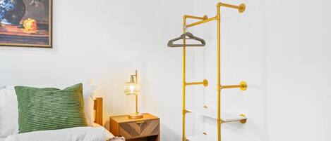 A bedroom with a bed, a bedside table with a lamp, a hanging clothes rack, and a painting on the wall.