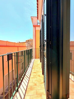 Un balcone stretto con ringhiera in metallo, pavimento piastrellato e vista sugli edifici vicini.