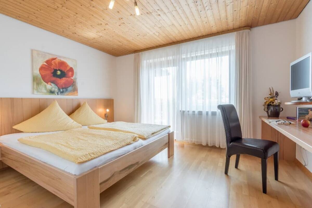 Une chambre à coucher avec un lit en bois, un bureau avec un ordinateur, une chaise et une fenêtre avec des rideaux.
