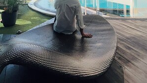 Un uomo seduto su una poltrona circolare in vimini accanto a una piscina con fondo in vetro e vista su un grattacielo.