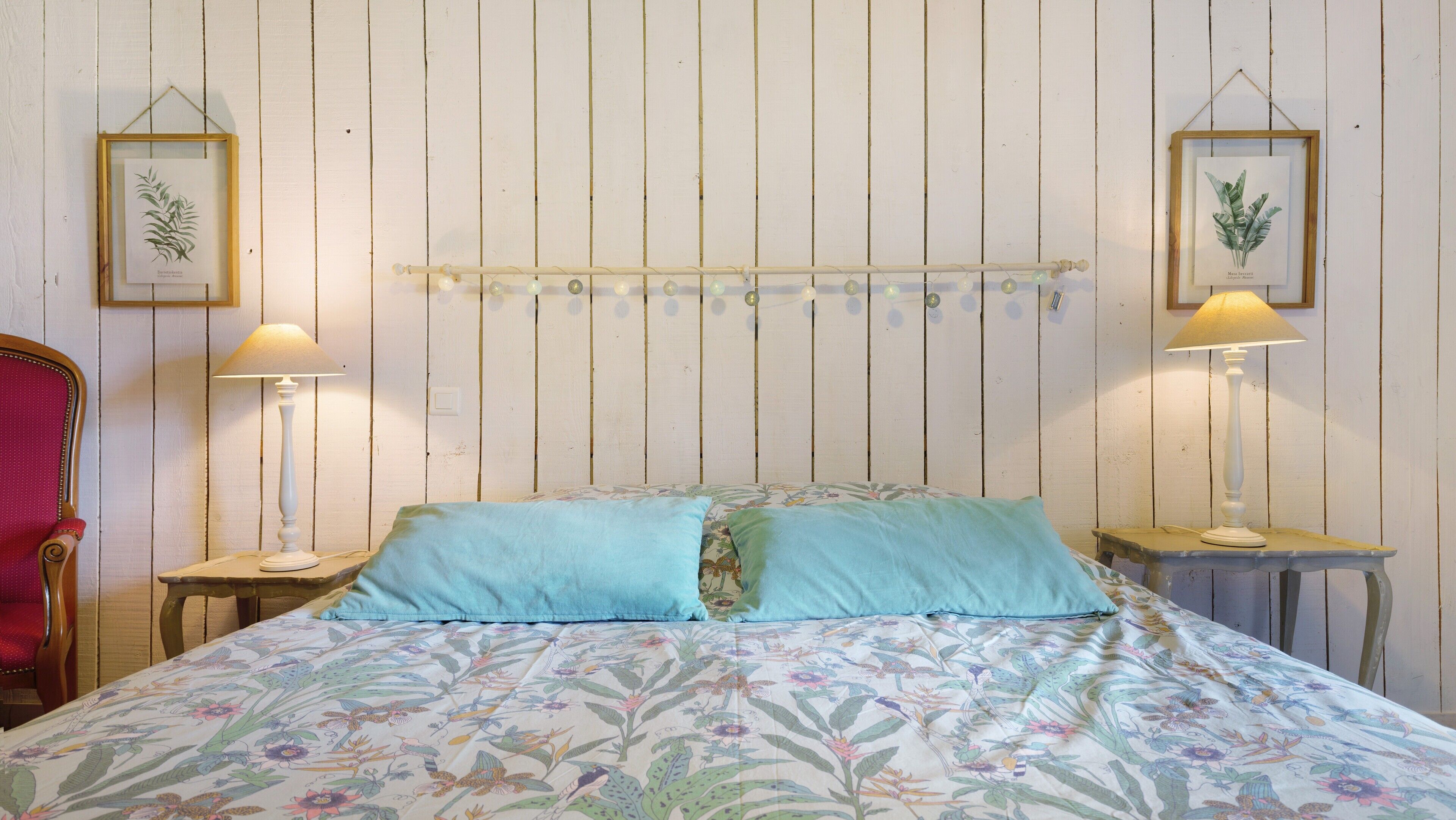 Ein Bett mit floraler Bettwäsche, zwei Nachttischlampen, eine Hängeleuchte und gerahmte Pflanzenbilder an den Wänden.