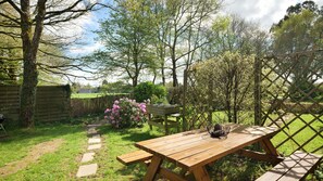 Ein Holztisch für Picknickzwecke mit Bänken, umgeben von einem Garten mit blühenden Pflanzen und Bäumen.