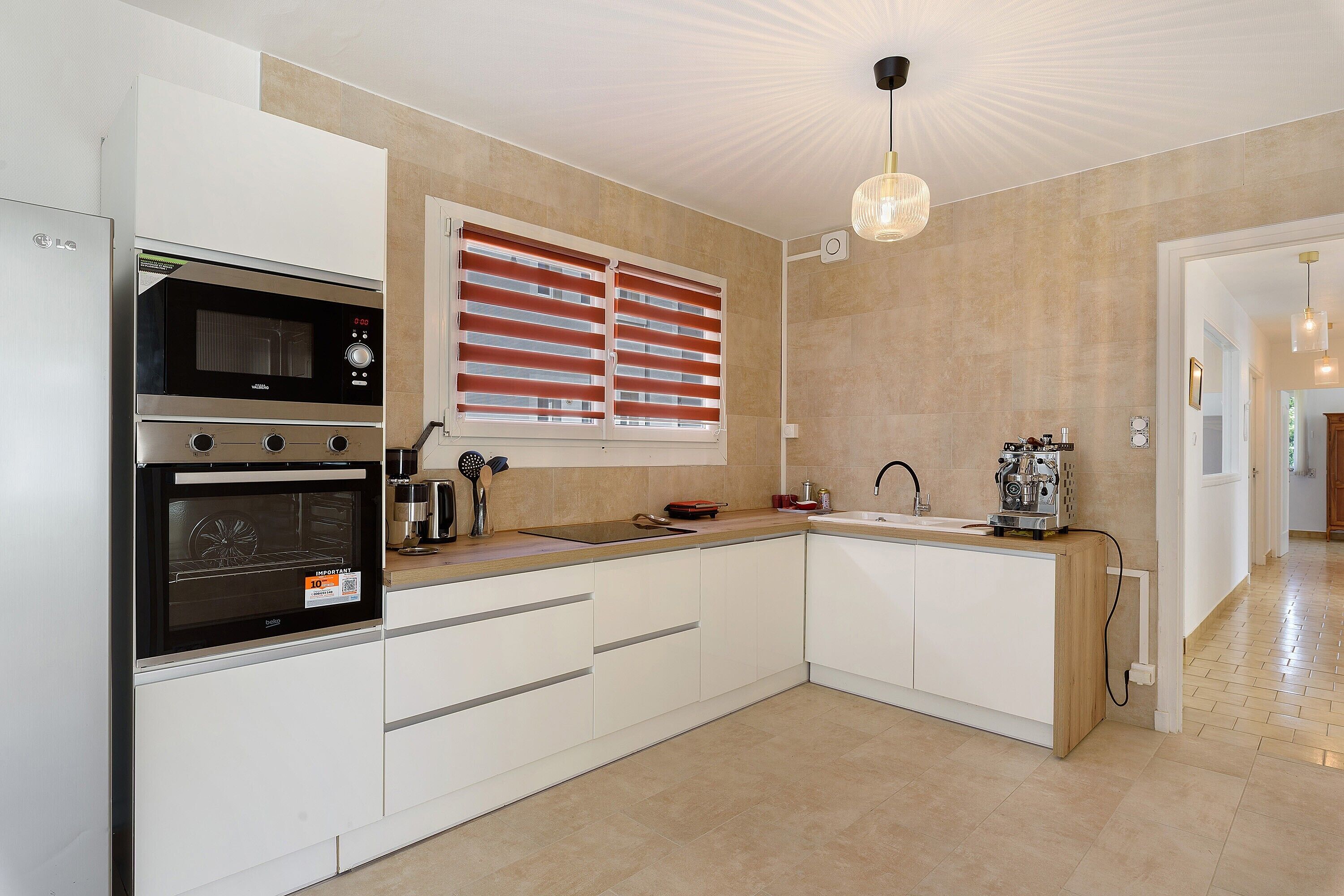 Une cuisine moderne dotée d’appareils électroménagers intégrés, d’une fenêtre avec stores et d’un luminaire suspendu.