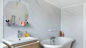 Un baño con lavamanos blanco, espejo y bañera. Hay artículos de aseo en el lavamanos y una toalla en el suelo.