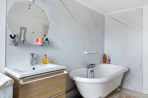 A bathroom with a white sink, mirror, and bathtub. There are toiletries on the sink and a towel on the floor.