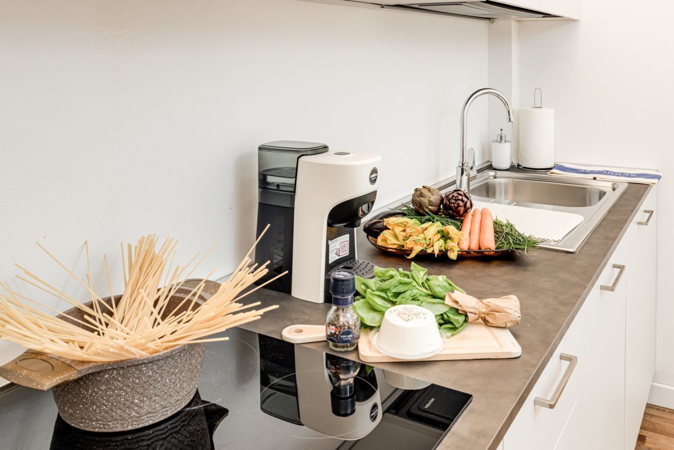 Une cuisine au design moderne, dotée d’un évier en acier inoxydable, d’une machine à café intégrée et d’une variété de légumes frais disposés sur le plan de travail.