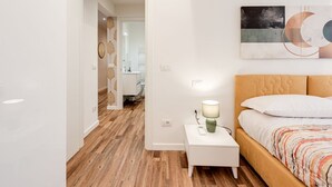 A bedroom with a wooden floor, a white nightstand with a lamp, a bed with a patterned bedspread, and a wall-mounted abstract painting. - Modern and cozy apartment, freshly renewed!. It features 2 fully equipped bathrooms with shower, WC, and bidet. The kitchen is brand new and fully equipped with an oven and microwave. The property includes 4 air conditioning and heating units, one in eac (Firenze)