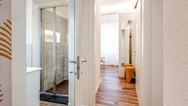 A hallway leading to a bathroom with a glass shower door, a wooden floor, and a small table with a chair. - Modern and cozy apartment, freshly renewed!. It features 2 fully equipped bathrooms with shower, WC, and bidet. The kitchen is brand new and fully equipped with an oven and microwave. The property includes 4 air conditioning and heating units, one in eac (Firenze)