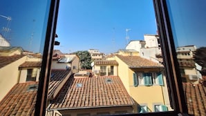 Vista pela janela de uma área residencial com telhados de telha de barro e céu azul límpido.