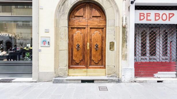Uma porta dupla de madeira com arco de pedra, uma loja com persiana vermelha e uma placa com o texto 'BE BOP'.