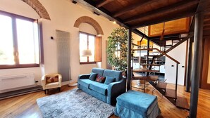 Sala de estar com um sofá azul, tapete cinza, piso de madeira, escada e uma planta.