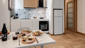 Una cocina con armarios blancos, salpicadero de azulejos, un refrigerador y una mesa de comedor con comida y bebidas.