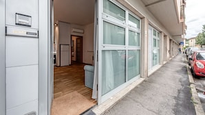 Entrada de un edificio con puerta de vidrio corredera, unidad de aire acondicionado visible y una pequeña zona de estar en el interior.