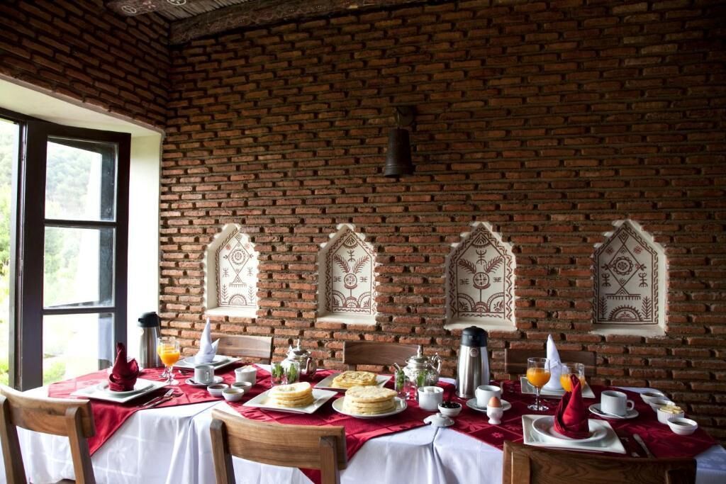 Comedor con pared de ladrillo, sillas de madera, una mesa dispuesta con platos, vasos y mantel rojo.