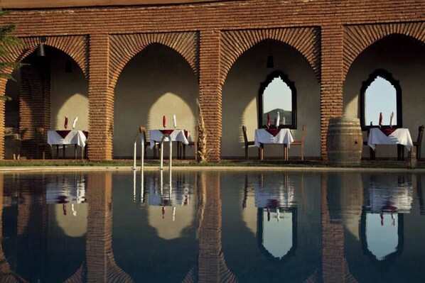 A pool with clear reflections, a brick structure with arches, and tables with white tablecloths.