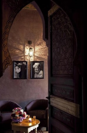 A dimly lit room with a decorative archway, a framed photograph, a table with a vase of flowers, and a patterned door.