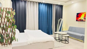 Hotellihuone, jossa on sänky, harmaa sohva, pieni pyöreä pöytä, kattotuuletin ja seinälle asennettu televisio, jossa näkyy kuva takkatulesta.