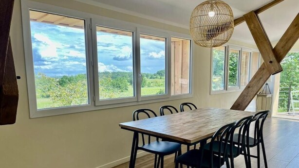 Un espace repas avec une table et des chaises en bois, de grandes fenêtres donnant sur un paysage verdoyant et un luminaire en fibres tressées.