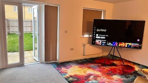 A room with a large flat-screen TV on a stand, a colorful rug, and a sliding glass door leading to a backyard. - House- Sleeps 6 & Free Parking (Grays)