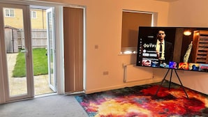 A room with a large flat-screen TV on a stand, a colorful carpet, a sliding glass door leading outside, and a small window. - House- Sleeps 6 & Free Parking (Grays)