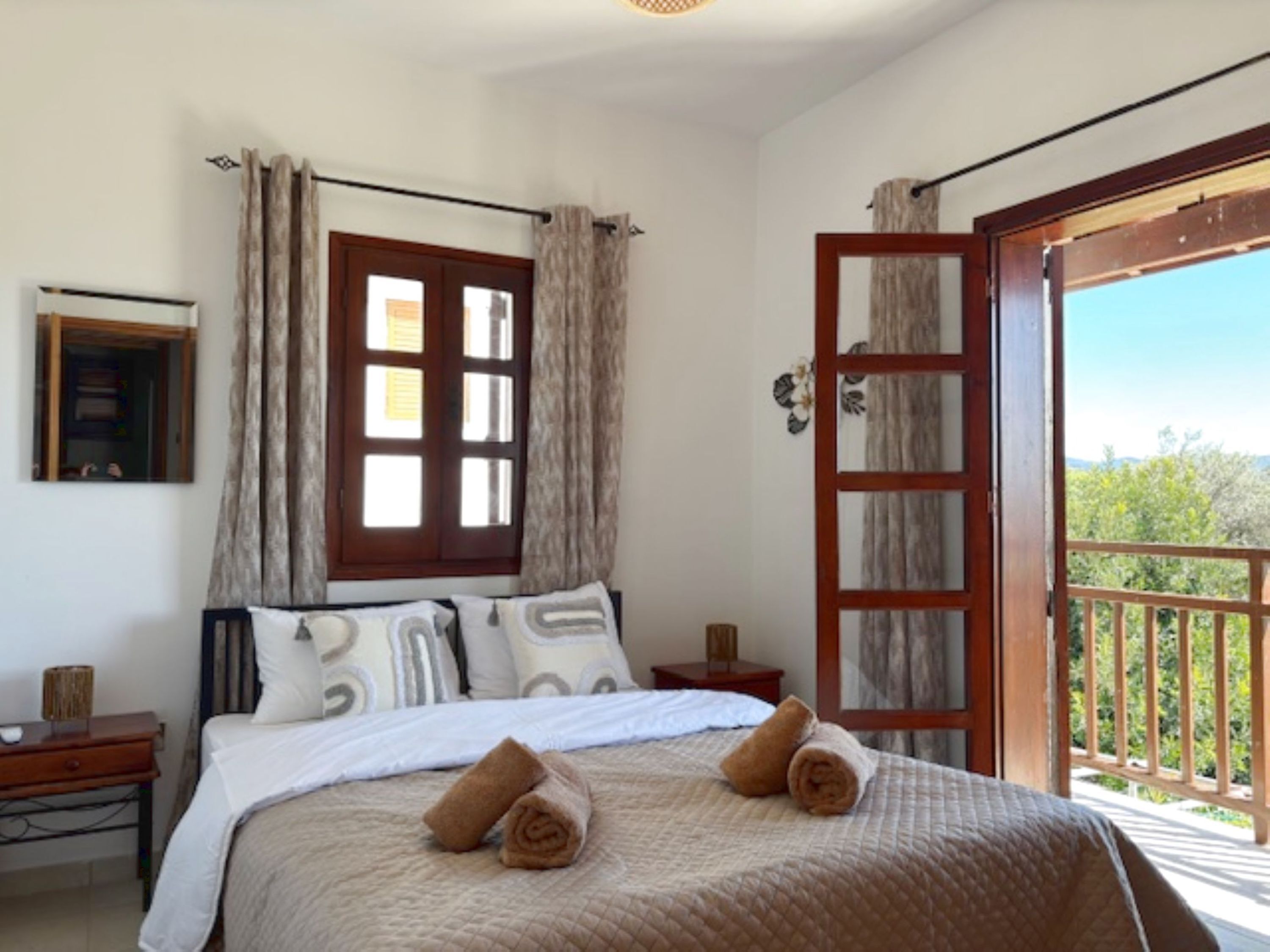 A bedroom with a bed, a nightstand, a window with curtains, a door leading to a balcony, and a framed picture on the wall.