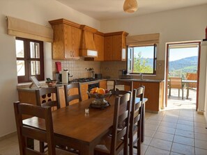 A kitchen with wooden cabinets, a dining table with chairs, a fruit bowl, and a view of the outdoors. - St Visianos Stone House (Filousa)