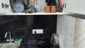 A kitchen with a black countertop, a coffee maker, a scale, and various kitchen items on the shelves. - Sunshine Stays, Gaur City Centre, Gr Noida West (Greater Noida)