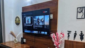 A reception desk with a flat-screen TV, a vase with flowers, and a wall-mounted light fixture. - Sunshine Stays, Gaur City Centre, Gr Noida West (Greater Noida)