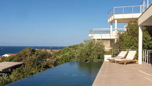 Una piscina a sfioro moderna con vista sull'oceano, un balcone con poltrone relax e vegetazione rigogliosa.