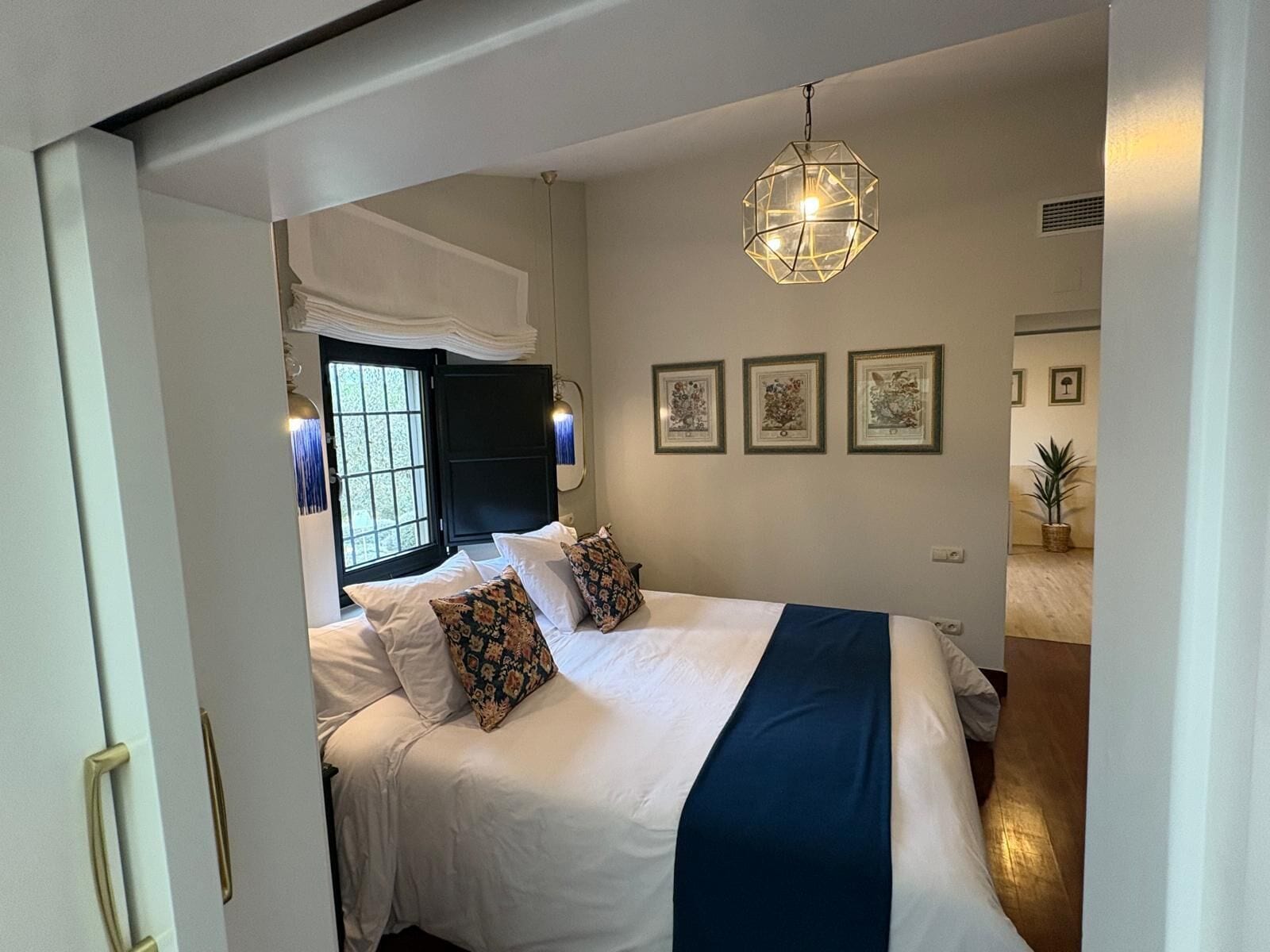 Ein Schlafzimmer mit einem Bett, einem Vorhangfenster, einer Hängeleuchte und gerahmten Bildern an der Wand.