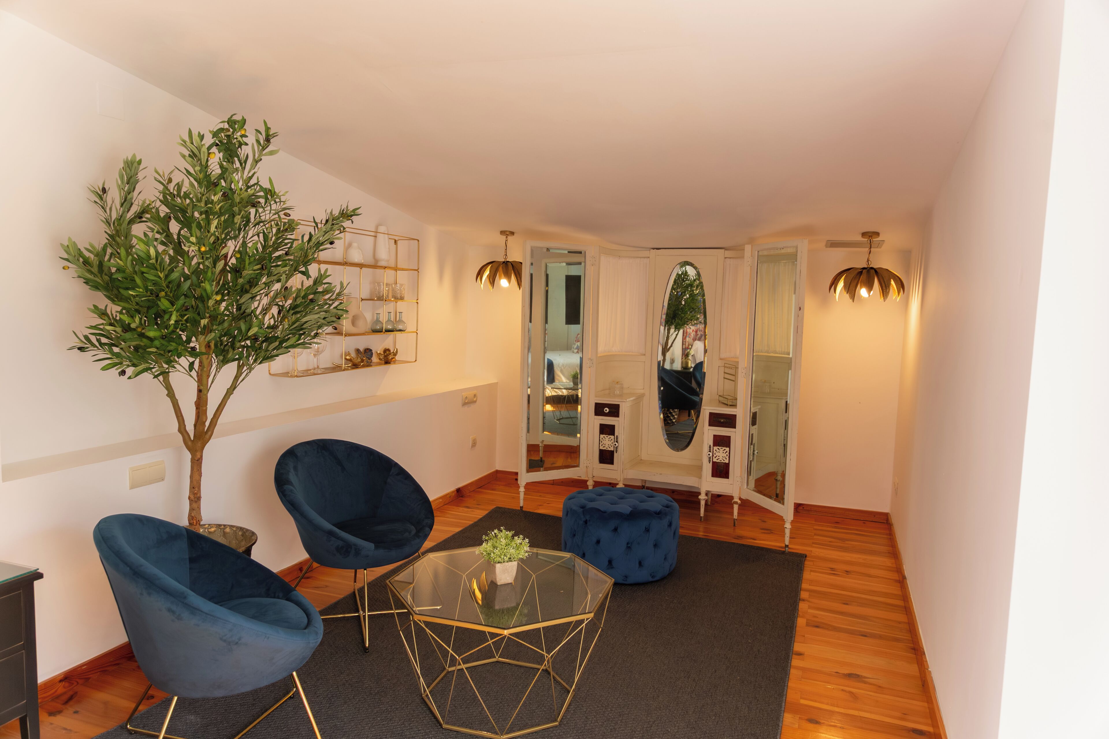 A modern living room with a glass coffee table, two blue armchairs, a small tree, and a mirror.