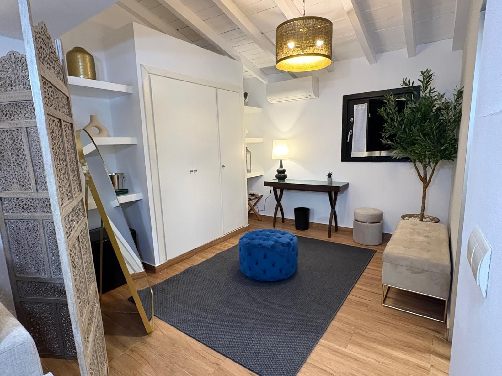 A modern living room with a large wardrobe, a dining table, a blue ottoman, a small potted plant, and a hanging light fixture.