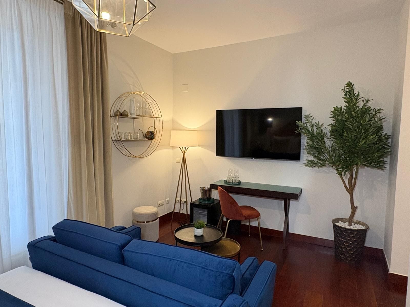 A modern living room with a blue sofa, a round coffee table, a TV mounted on the wall, a small desk, and a potted plant.