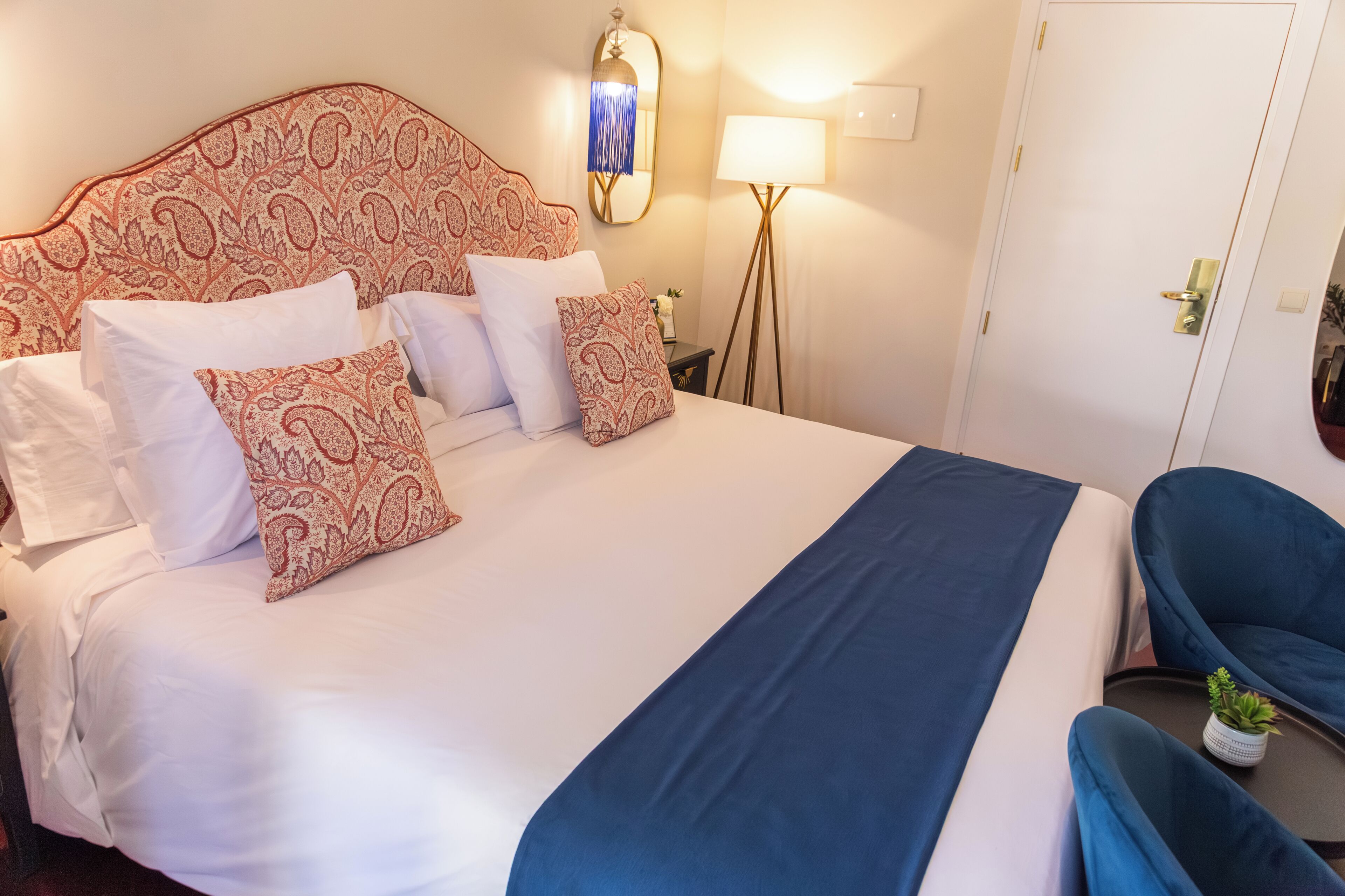 A neatly made bed with patterned pillows, a bedside lamp, and a chair with a plant.