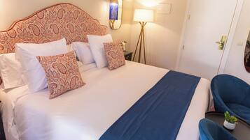 Een netjes opgemaakt bed met patroon kussens, een bedlampje en een stoel met een plant.